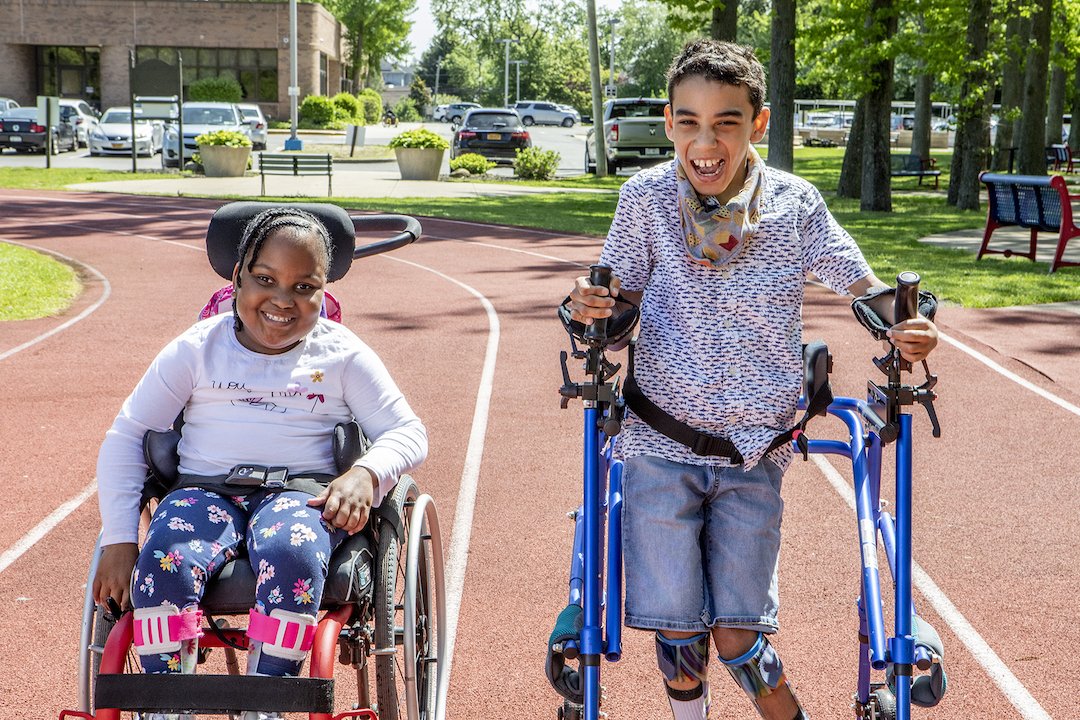 Henry Viscardi School Hero Image - two students on a clay track, one in a wheelchair and one in a walker both smiling