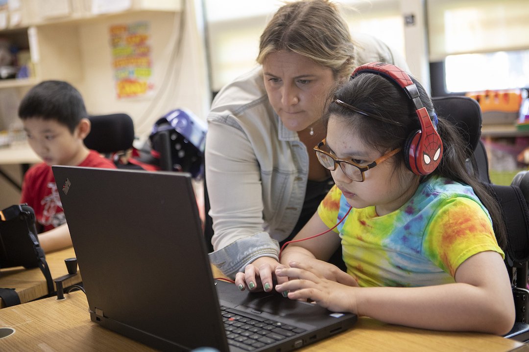 Henry Viscardi School - Join our diverse team image with a teacher reaching over the shoulder of a young student with headphones in a wheelchair to help them with their schoolwork on a laptop computer